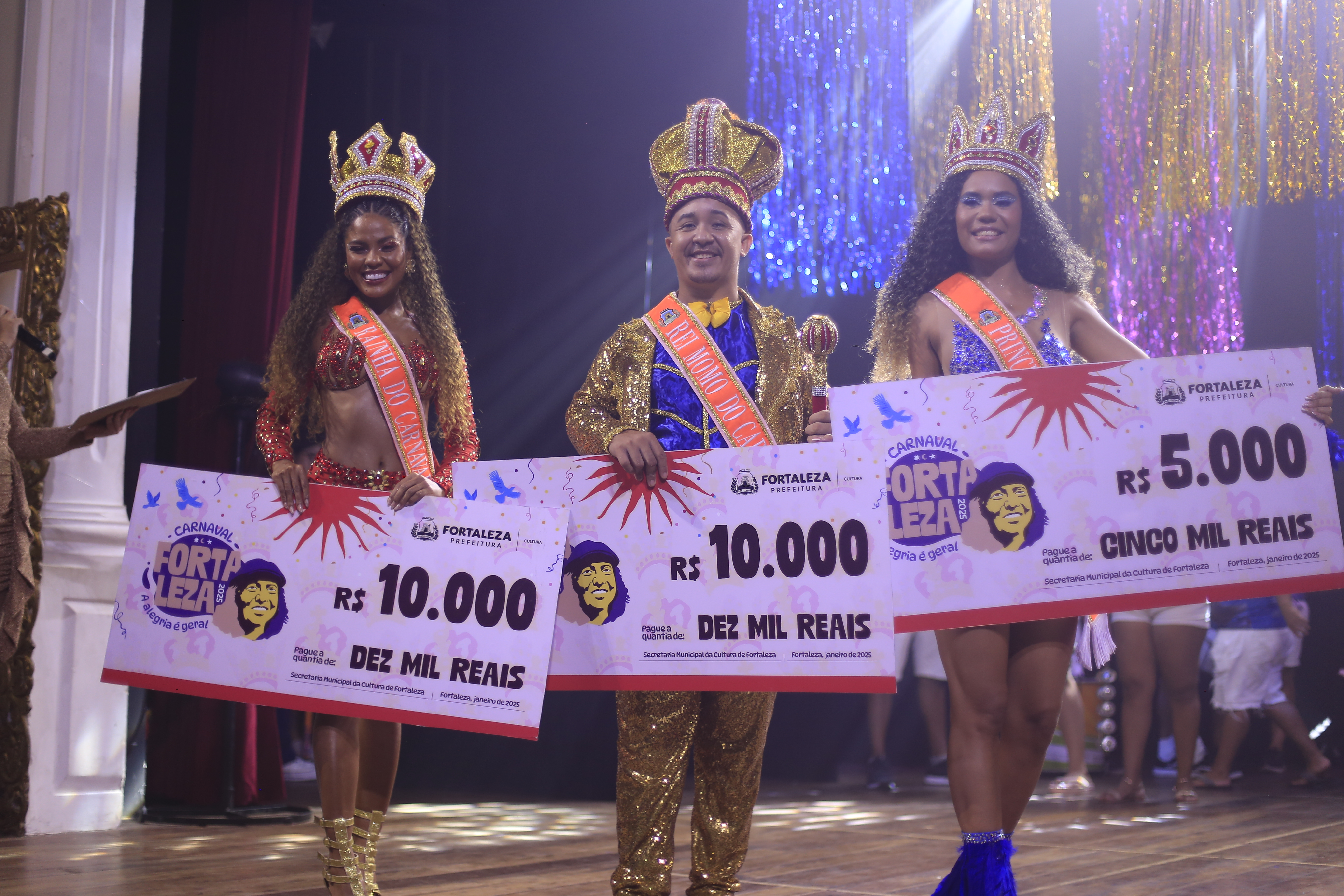 Rainha Cláudia Gonçalves da Conceição, Rei Jonathan Silvino da Silva e Princesa Ana Beatriz Feijó Mendes posam para a foto no palco do teatro são josé, segurando os cheques pela premiação